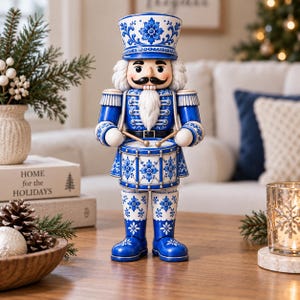 May include: A nutcracker figurine featuring a blue and white floral pattern on its uniform, hat, and drum. The figure has a white beard, a black mustache, and holds two drumsticks. It is dressed in a blue jacket with silver accents.