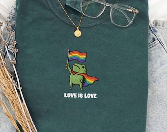 Könnte beinhalten: Dunkelgrünes T-Shirt mit einer gestickten Froschfigur, die eine Regenbogenflagge hält und einen Regenbogenumhang trägt. Der Text "LOVE IS LOVE" steht unter dem Frosch. Eine goldene Halskette und eine klare Brille liegen auf dem Shirt.