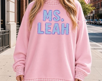 Könnte beinhalten: Ein hellrosa Sweatshirt mit hohem Kragen, Bündchen und einem unteren Saum mit rosa-weißem Vichy-Muster. In der Mitte der Brust befindet sich der Text MS. LEAH in blau-weiß karierten Blockbuchstaben mit einer violetten gestickten Umrandung.