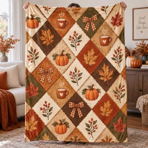 May include: A soft throw blanket featuring a diamond-patterned design with alternating squares of cream and autumn-toned colors. The print includes illustrations of orange pumpkins, brown coffee mugs, red berries, maple leaves, and plaid bows.