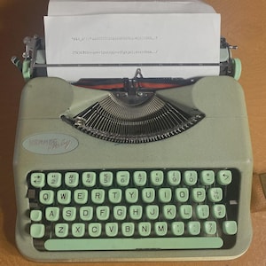 May include: A vintage Hermes Baby typewriter in a pale green hue. The keyboard features light green keys with white lettering. A sheet of paper with typed text is inserted. The typewriter rests on a brown surface.