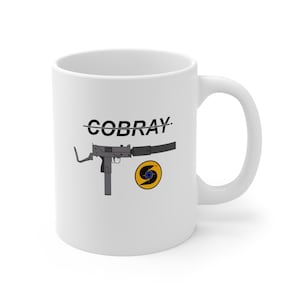 May include: White ceramic coffee mug with a black and white illustration of a submachine gun and the text "COBRAY" with a line through it. A yellow and blue target symbol is below the gun.