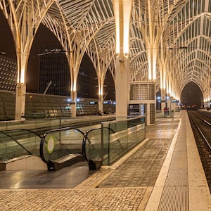 May include: A modern train station with a white metal vaulted ceiling and a long platform with train tracks. The station is lit with warm yellow lights.