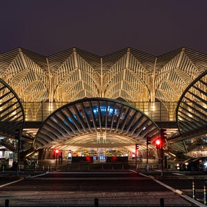 May include: A modern, illuminated train station with a large, arched glass roof and a metal framework. The station is lit up at night, and there are cars driving on the road in front of it.