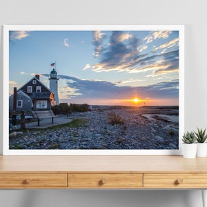 May include: A white lighthouse with a red roof and a small house next to it. The lighthouse is on a rocky beach with a cloudy sky and the sun setting in the distance.