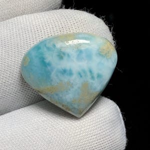 May include: A polished, heart-shaped gemstone with a light blue and white pattern, accented with tan patches. The stone is held against a white fabric background, showcasing its natural beauty and unique color variations.