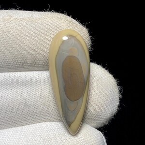 May include: A polished, teardrop-shaped gemstone with a layered pattern of tan, gray, and brown hues. The stone is held against a white fabric background, showcasing its smooth, reflective surface. The gemstone's elongated form is ideal for jewelry.