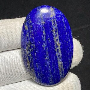 Könnte beinhalten: Ovaler Lapis Lazuli-Edelstein. Der polierte Stein hat eine tiefblaue Farbe mit goldenen und weißen Streifen. Der Edelstein wird in einer weißen behandschuhten Hand gehalten.