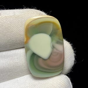 May include: A polished, rectangular gemstone with a swirling pattern of colors. The stone features a central, creamy white shape surrounded by bands of green, brown, and yellow. The gemstone is held by a white gloved hand.