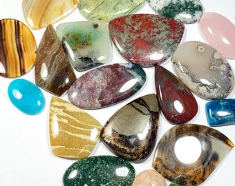 May include: A collection of polished gemstones in various shapes and colors, including brown, green, red, and blue. The gemstones are displayed on a white surface, showcasing their unique patterns and textures. The collection includes oval, teardrop, and rectangular shapes.