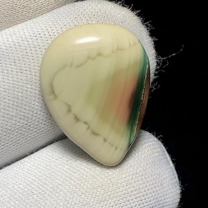 May include: A polished, teardrop-shaped gemstone with a creamy white base color. The stone features bands of light green, pink, and brown hues, creating a unique, natural pattern. The gemstone is held against a white fabric background.