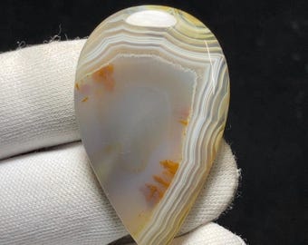 May include: A teardrop-shaped agate stone with a polished surface. The stone features layers of white, tan, and yellow hues, with orange and brown inclusions. The agate is held by a gloved hand, showcasing its natural patterns.