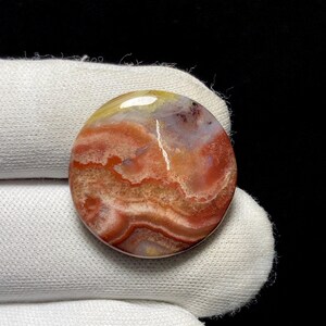 Puede incluir: Una gema redonda y pulida con un patrón arremolinado de tonos rojos, naranjas, amarillos y blancos. La piedra tiene una superficie lisa y brillante y parece estar sujeta por un guante blanco. Producto natural.