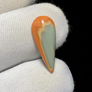 May include: A teardrop-shaped gemstone with a polished surface. The stone features a blend of colors, including orange, cream, and pale green. The gemstone is held against a white fabric background, with a black background.
