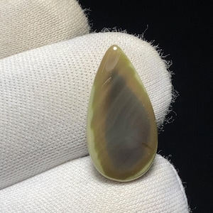 May include: A teardrop-shaped gemstone with a polished surface. The stone features a blend of colors, including light green, brown, and a darker grey-brown in the center. The gemstone is held against a white fabric background.