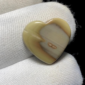 May include: A heart-shaped stone with a polished surface. The stone features a blend of beige, cream, and brown hues, with a subtle pattern. The heart is smooth and appears to be a natural gemstone, possibly for jewelry or crafts.
