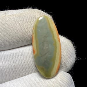 May include: An oval-shaped gemstone with a polished surface. The stone features a blend of colors, including gray, yellow, orange, and green, creating a striped pattern. The gemstone is held in a white fabric.