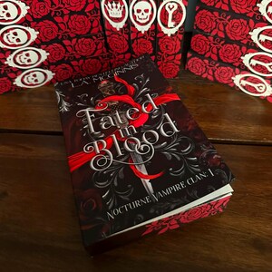 May include: A hardcover book titled "Fated in Blood" with a dark cover featuring a sword and red ribbon design. The book is part of the "Nocturne Vampire Clan" series. Decorative bookmarks with skulls, crowns, and keys are in the background.