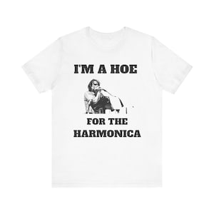 May include: A white short-sleeve t-shirt featuring a black and white graphic of a person playing the harmonica into a microphone. The text printed in black block letters above and below the image reads, I'M A HOE FOR THE HARMONICA.