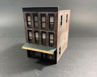Może przedstawiać: Miniaturowy ceglany budynek z czarnym dachem i ramami okiennymi. Budynek ma dwa piętra z wieloma oknami i zieloną markizą z napisem "FLOWERS BY TERRY".