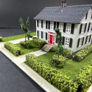 May include: A miniature white two-story house with a black roof, black shutters, and a red front door. The house is surrounded by a green lawn, hedges, and a sidewalk, creating a detailed model of a residential property.