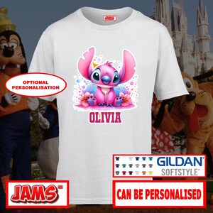 May include: White t-shirt featuring a pink cartoon character with large ears and the name "OLIVIA" in pink. The design includes blue and pink accents. The shirt has a Gildan Softstyle tag.