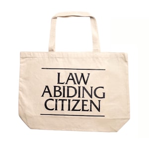Può includere: Una borsa tote in tela beige con manici neri e il testo "LAW ABIDING CITIZEN" stampato in nero.