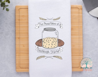 May include: White kitchen towel with a brown and white illustration of a jar of sourdough starter and two loaves of bread. The text reads "The Proud Parent of a Sourdough Starter".