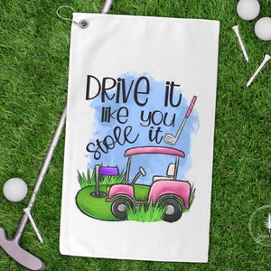 May include: White golf towel with the text "Drive it like you stole it" and a pink golf cart graphic. The towel also features a golf club, golf ball, and tee on a green grass background.