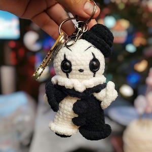 May include: A handmade crochet keychain featuring a small, whimsical jester doll. The doll has a white body with black accents, including a hat, sleeves, and facial details. It has large, black button eyes and a ruffled collar. The keychain includes a metal ring and keys.