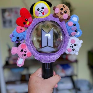 May include: A black and purple light stick with a clear orb featuring a silver logo. The orb is surrounded by a plush purple ring adorned with small, colorful, crocheted character figures. The light stick is held in a hand.