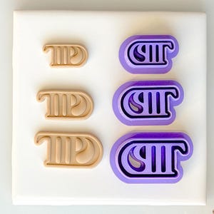 Peut inclure: Ensemble de six emporte-pièces, trois beiges et trois violets, chacun avec un motif de lettre stylisé. Les emporte-pièces sont disposés sur une surface blanche. Une cloche et un ruban dorés sont en arrière-plan.