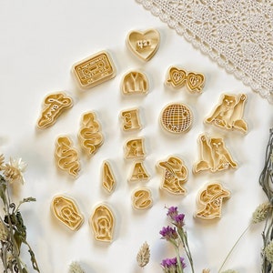 May include: A set of 18 cookie cutters in various shapes, including a heart, a disco ball, a cassette tape, a cowboy boot, a snake, a crystal, sunglasses, a jacket, and the letters "ERA". The cutters are made of a light beige plastic and are arranged on a white surface.