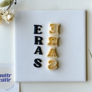 May include: A set of black and white plastic cookie cutters in the shape of the letters "ERAS". The cutters are arranged vertically on a white surface.