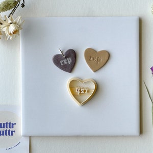 May include: Three heart-shaped cookie cutters, one brown, one beige, and one white. The cutters are on a white surface. The brown and beige cutters have the word "Mom" written on them. The white cutter has the word "Dad" written on it. The cutters are for making cookies or other baked goods.