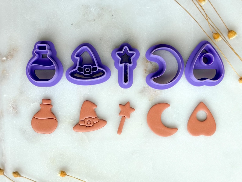 May include: A collection of purple and orange Halloween-themed cookie cutters. The shapes include a potion bottle, witch hat, star wand, crescent moon, and a planchette. The cutters are arranged in two rows, with the purple cutters on top and the orange cutters below.
