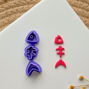 May include: Two sets of fish-shaped cookie cutters. One set is purple and the other is red. The purple set has three pieces: a fish head, a fish body, and a fish tail. The red set has three pieces: a fish head, a fish body, and a fish tail.