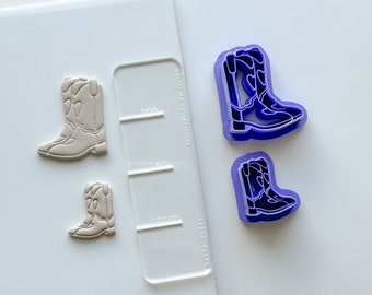 Puede incluir: Tres cortadores de galletas con forma de botas de vaquero. Dos son de plástico morado y uno es beige. El cortador beige está sobre una superficie blanca con una guía de plástico transparente con las palabras "one", "two" y "three" impresas.