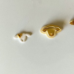 May include: Two sheep-themed items are shown against a white background. One is a small, textured, white sheep figurine with a yellow bow. The other is a beige sheep-shaped cookie cutter. These items are likely for baking or crafting.