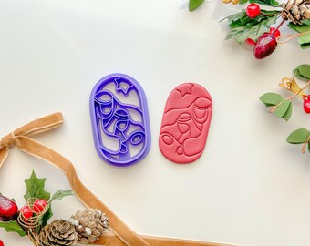 May include: Two Christmas cookie cutters, one purple and one red. The purple cutter is shaped like a star and has a design of three bells. The red cutter is shaped like a star and has a design of three figures.