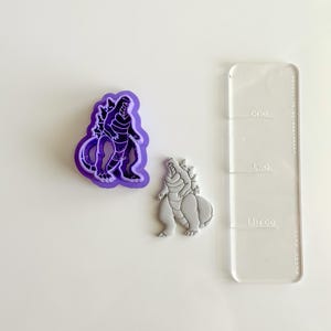 Puede incluir: Un cortador de galletas con forma de Godzilla morado y una galleta gris, junto a una regla acrílica transparente con las palabras "uno", "dos" y "tres". El cortador de galletas tiene un contorno morado.