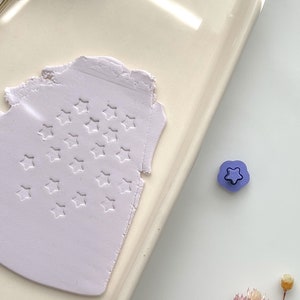 May include: A close-up of a light purple polymer clay with a star-shaped design. A small purple star-shaped stamp is visible in the background.