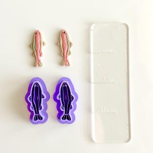May include: An overhead shot shows two fish-shaped cookie cutters in purple, two fish-shaped clay figures, and a clear acrylic ruler. The clay fish are pink, tan, and green. The ruler has the words "one", "two", and "three" printed on it.