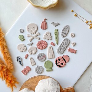 May include: A collection of Halloween-themed clay ornaments, including ghosts, pumpkins, bats, skulls, and candy corn. The ornaments are arranged on a white surface with a wooden tray holding two white pumpkins in the foreground.