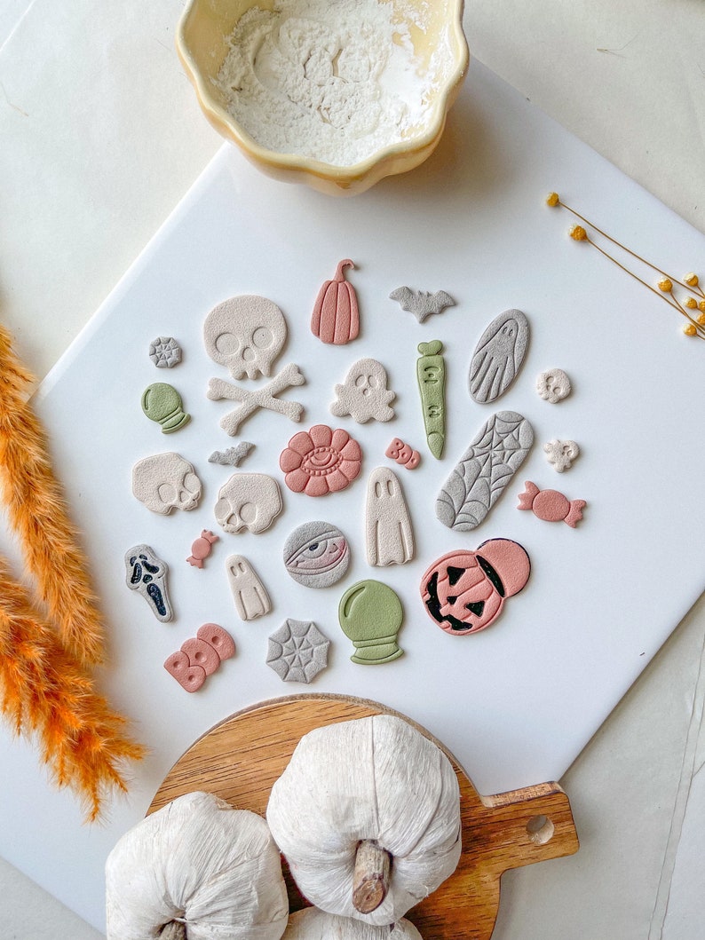 May include: A collection of Halloween-themed clay ornaments, including ghosts, pumpkins, bats, skulls, and candy corn. The ornaments are arranged on a white surface with a wooden tray holding two white pumpkins in the foreground.