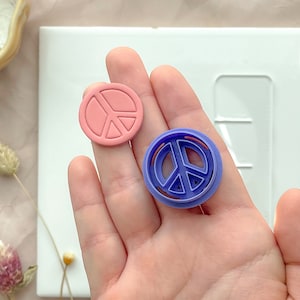 Può includere: Due tagliapasta a forma di segno della pace, uno rosa e uno viola, tenuti in una mano.
