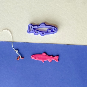 Peut inclure: Un emporte-pièce en forme de poisson violet et un biscuit en forme de poisson rose sur un fond bleu. Une ficelle blanche avec un crochet rouge pend du haut de l'image.