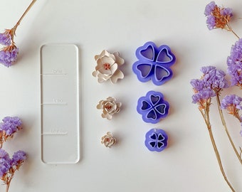 May include: Five purple plastic flower-shaped cookie cutters of varying sizes, arranged vertically on a white surface. The cutters are labeled "one", "two", and "three" on a clear plastic ruler to the left.