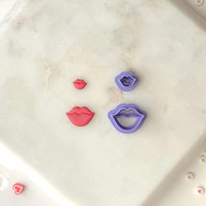 Pode incluir: Quatro cortadores de biscoitos em forma de lábios, em vermelho e roxo. Dois lábios vermelhos e dois cortadores roxos em forma de lábios são exibidos em uma superfície de mármore branco. Os cortadores variam em tamanho.