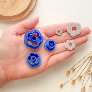 Puede incluir: Un juego de cinco cortadores de galletas de plástico en forma de flor, azules y grises. Los cortadores están dispuestos en una mano, con el cortador más grande en el centro y el más pequeño a la derecha.
